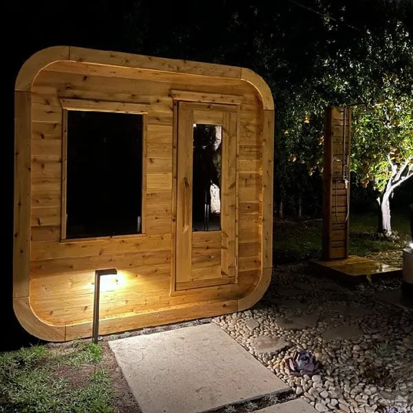 Cedar cube sauna at night lit by a garden uplight, front window and glass-panel door, timber outdoor shower post and pebble garden beside.