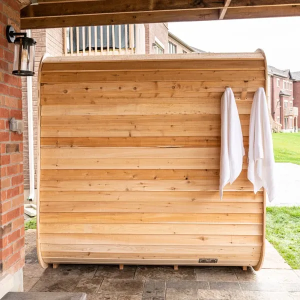 Side wall of a cube sauna in horizontal light cedar boards with towels on hooks, under a covered patio beside a brick house.