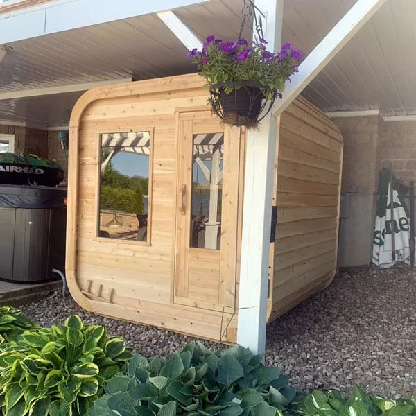 Cedar cube sauna with front window and glass-panel door on gravel under a covered patio, hanging flower basket in front, hot tub beside.