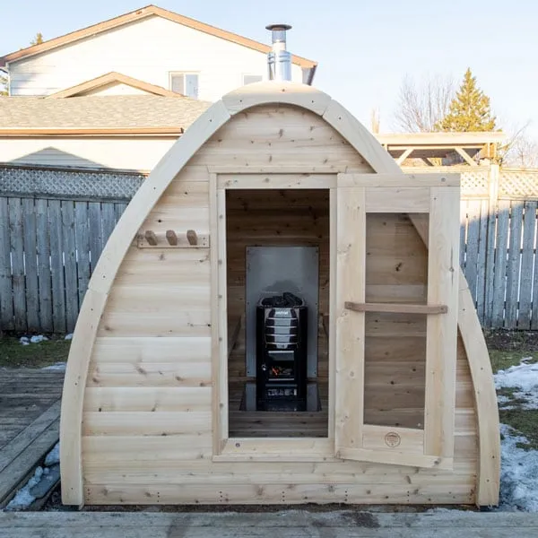 Front of an arched cedar pod sauna with door open, showing a wood-burning stove with stones against a steel heat shield; chimney above, backyard fence behind.