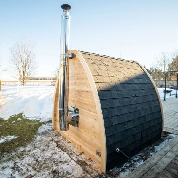 Rear three-quarter view of an arched pod sauna with dark shingle roof and light cedar end wall, stainless chimney exiting the rear wall, snowy yard beside a deck.