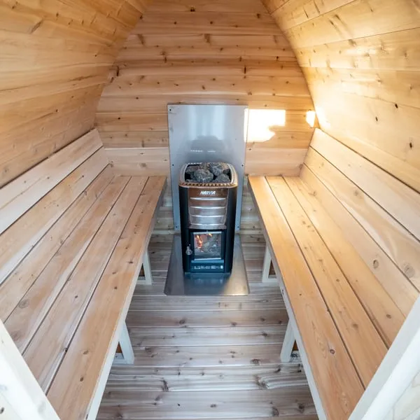 Pod sauna interior in cedar with benches along both sides and a wood-burning stove with stones against a steel heat shield on the rear wall.