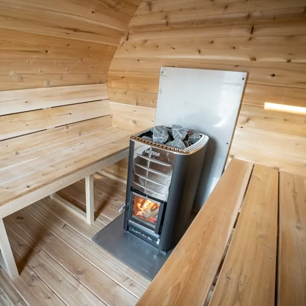 Close view of a wood-burning sauna stove with fire visible, stones on top, steel heat shield and floor plate, between cedar benches.