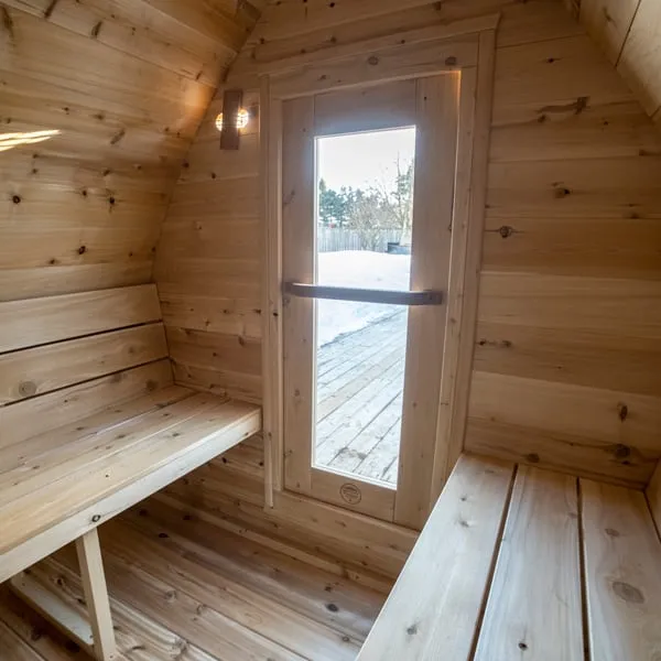 Cedar pod sauna interior looking toward the glass-panel door with wooden handle, snowy deck outside, side benches and wall light.