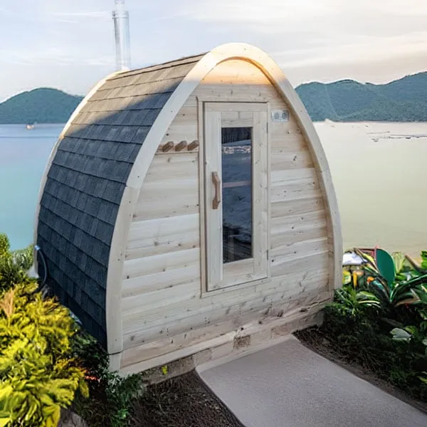 Arched pod sauna with dark shingle roof, cedar front wall, glass-panel door and chimney, beside a path above a bay with hills (appears composited).