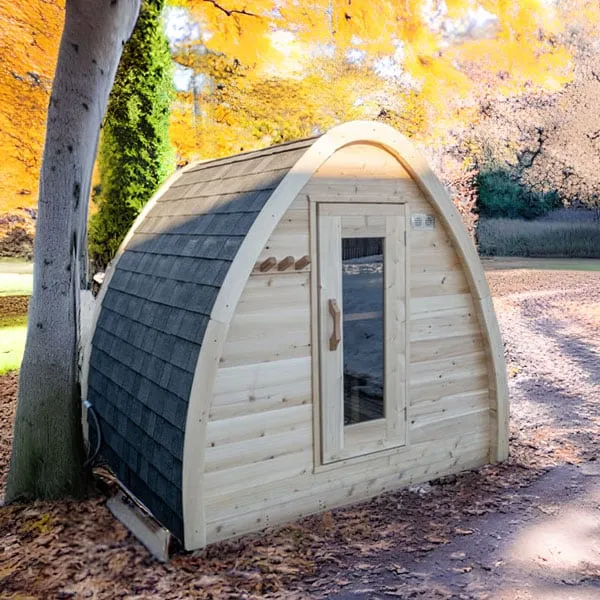Arched pod sauna with dark shingle roof, cedar front wall and glass-panel door, under a tree on leaf-covered ground in autumn (appears composited).