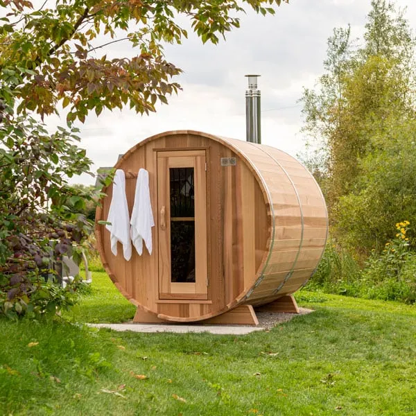 Barrel sauna in light cedar with glass-panel door, towels on hooks, steel bands and metal chimney, on a lawn among trees