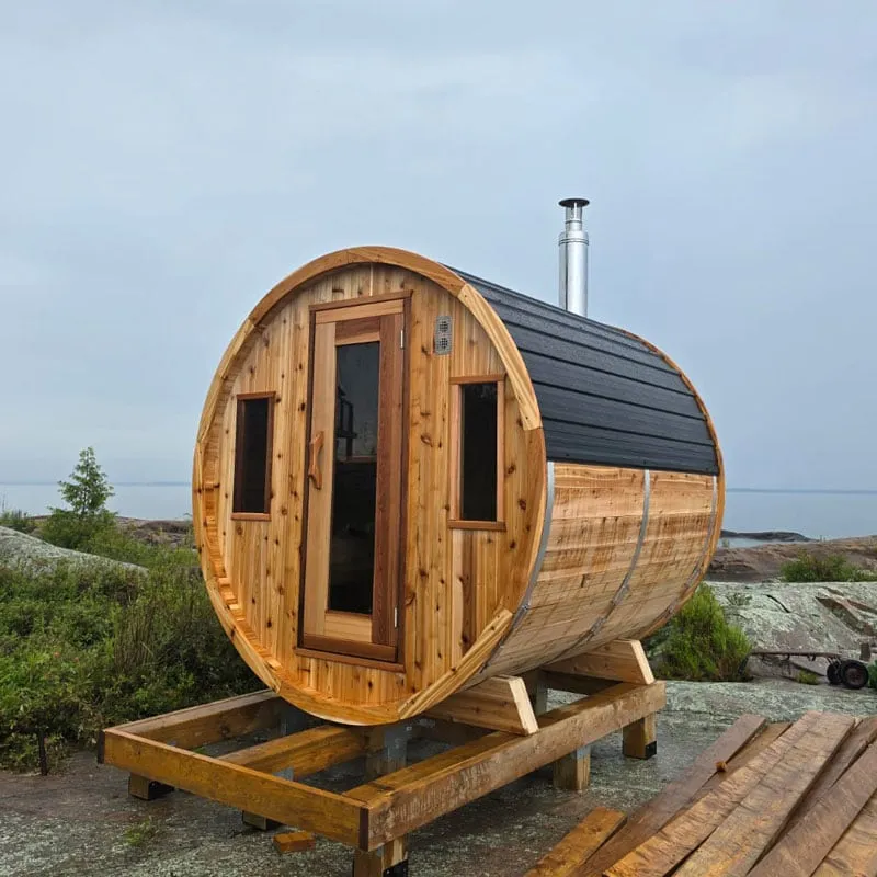 Knotty cedar barrel sauna with black shingle roof cover, glass door, two small front windows and chimney, on a timber base by a lake
