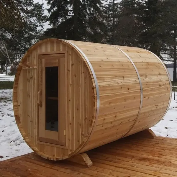 Knotty cedar barrel sauna with glass-panel door and steel bands on a timber deck, snowy yard with evergreens