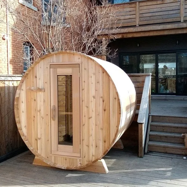 Light cedar barrel sauna with glass-panel door on a backyard deck beside a brick house and stairs