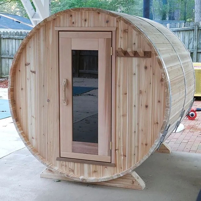 Knotty cedar barrel sauna with full-height glass door and towel pegs on a concrete patio, fence behind