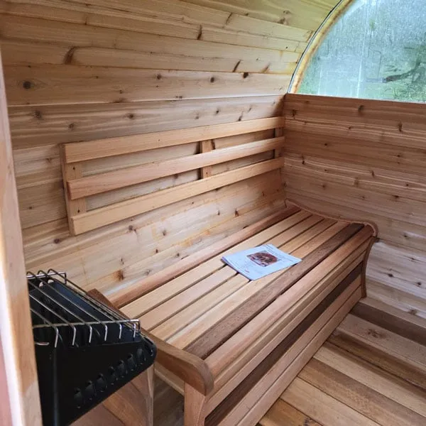 Barrel sauna interior: cedar bench with backrest, black electric heater with rock cage in foreground, half-moon end window