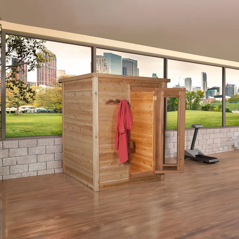 Indoor cedar cabin sauna with flat top, open framed glass door and red robe on wall pegs, shown in a gym room with city-view windows