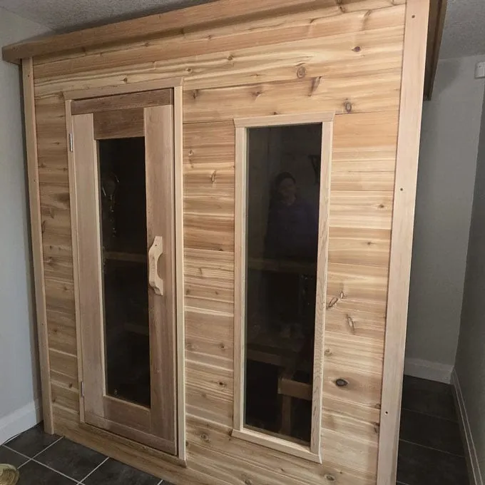Indoor cedar cabin sauna with framed glass door and tall side window, installed in a tiled basement room