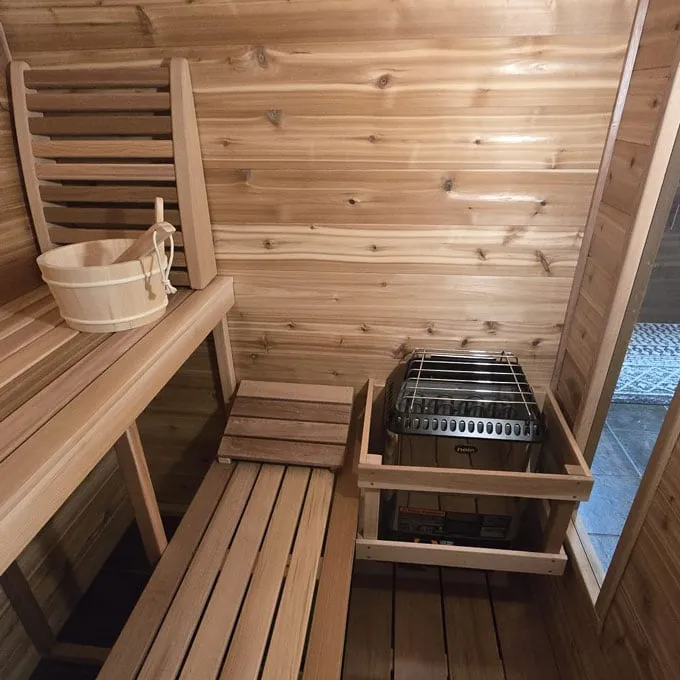 Cedar sauna interior with two-tier slatted benches, backrest, wooden bucket and ladle, and electric heater inside a timber guard