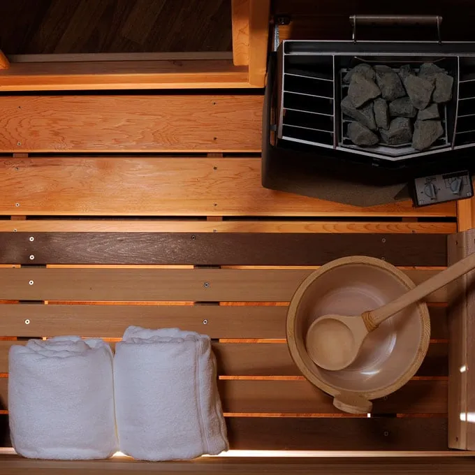 Overhead view of cedar sauna bench with rolled white towels, bucket and ladle beside an electric heater filled with stones and control knobs