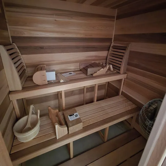 Cedar sauna interior with two-tier benches, two backrests, accessories in boxes, bucket and ladle, and a round caged electric heater in the corner