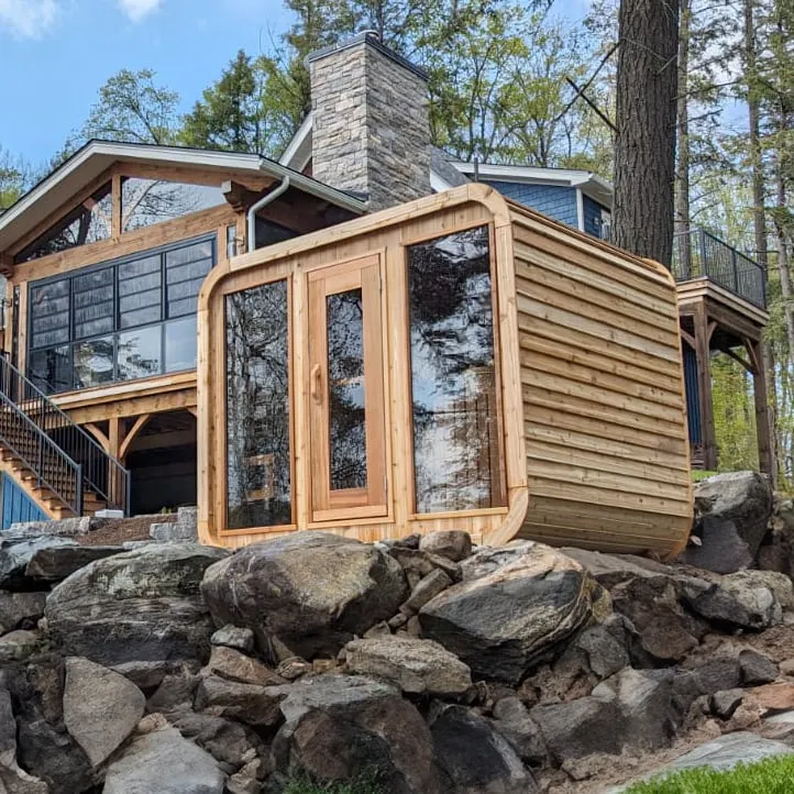 Rounded-corner cube sauna in cedar with glass door and two full-height windows on a rocky bank below a lake house