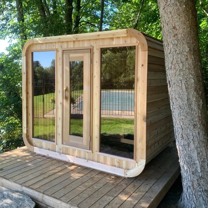 Cedar cube sauna with rounded corners, glass door and two large windows on a timber deck under a tree, pool behind