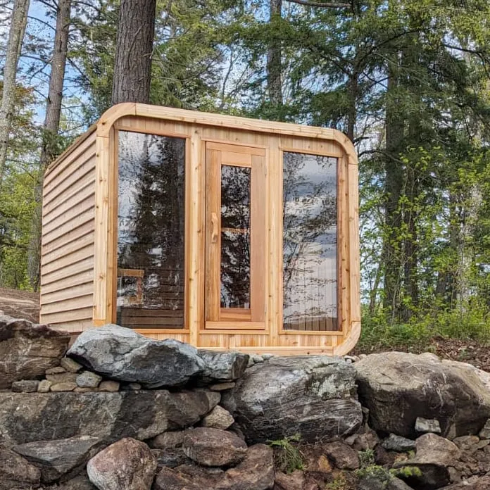 Cedar cube sauna with rounded corners, glass door and two tall windows above a stone wall, forest behind
