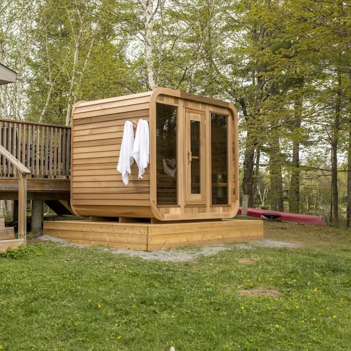 Cedar cube sauna with glass front and towels on a peg rack, on a raised timber platform in a wooded yard