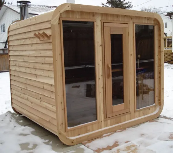 Cedar cube sauna with rounded corners, glass door, two large windows and metal chimney on a snowy patio