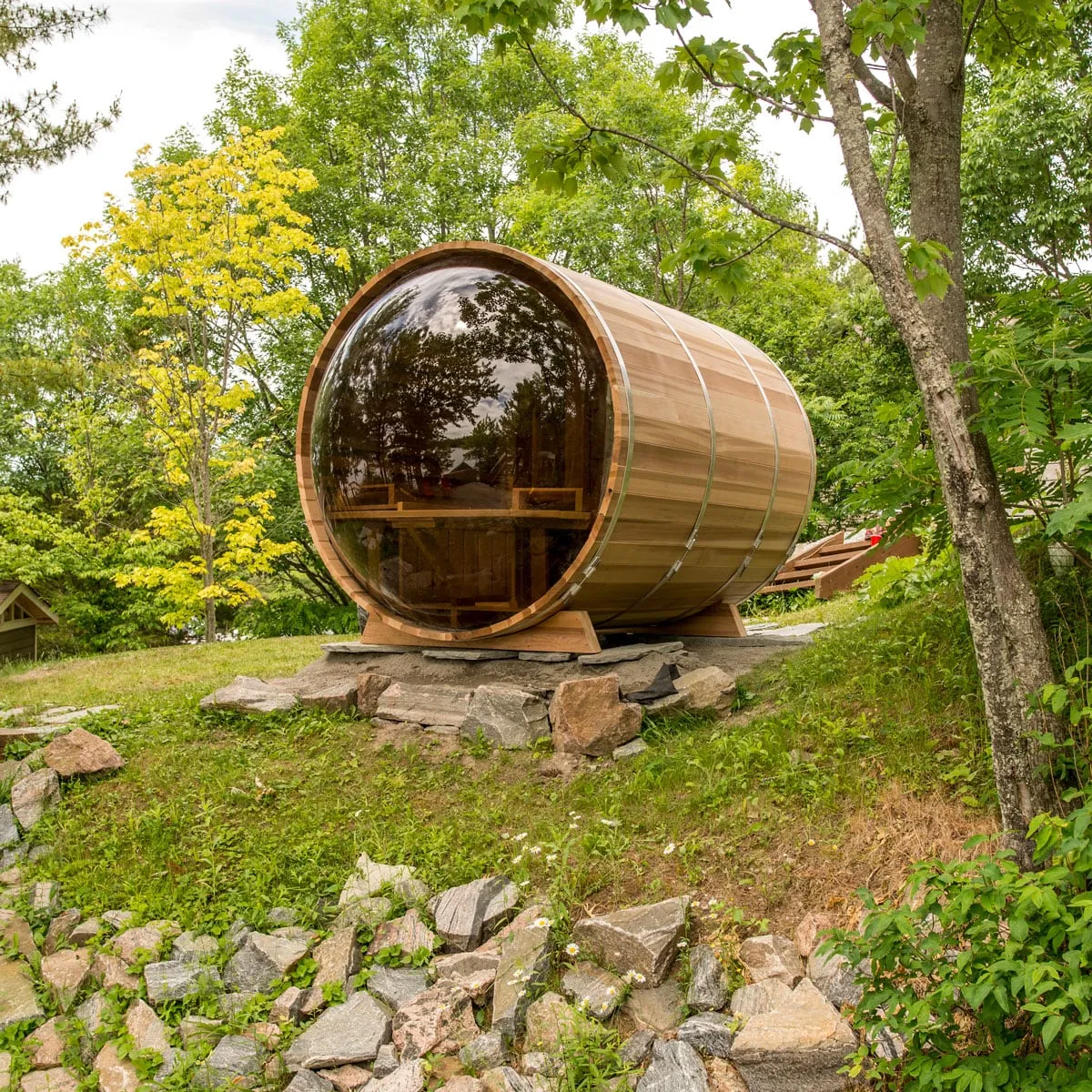 Cedar barrel sauna with full curved tinted glass end and steel bands, on a stone pad on a grassy slope among trees