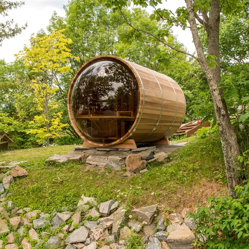 Cedar barrel sauna with full curved tinted glass end and steel bands, on a stone pad on a grassy slope among trees