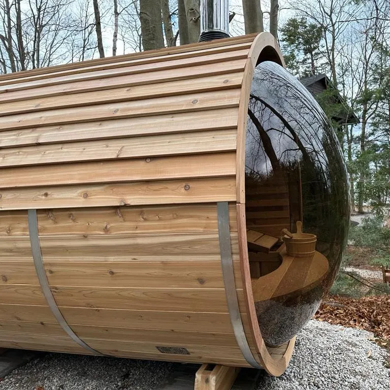 Knotty cedar panoramic barrel sauna, side view, with tinted curved glass end and chimney, on gravel in a wooded yard