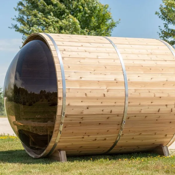 Knotty cedar barrel sauna with tinted curved glass end and steel bands on a lawn under blue sky