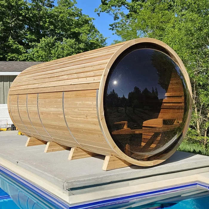 Long knotty cedar panoramic barrel sauna with tinted glass end on timber cradles on a pool deck, trees behind