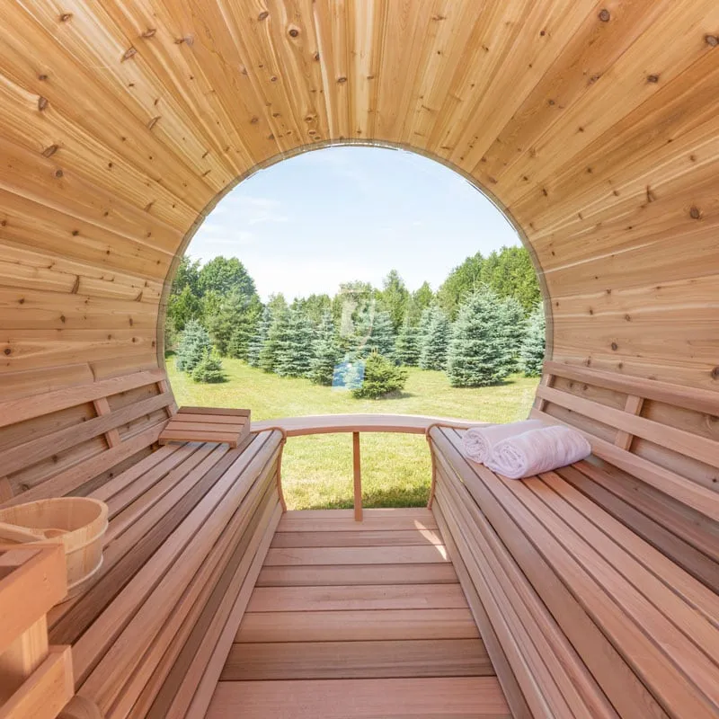 Barrel sauna interior looking out through the curved glass end: cedar benches both sides, towels, bucket, lawn and spruces