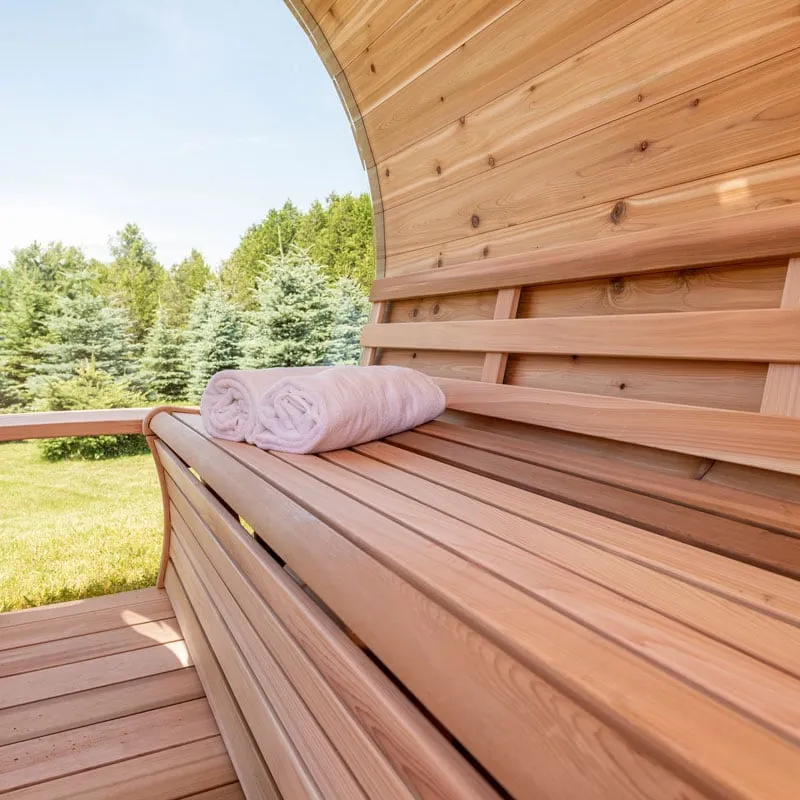 Cedar bench with backrest and rolled towels inside a barrel sauna, curved armrest, view through glass end to spruces