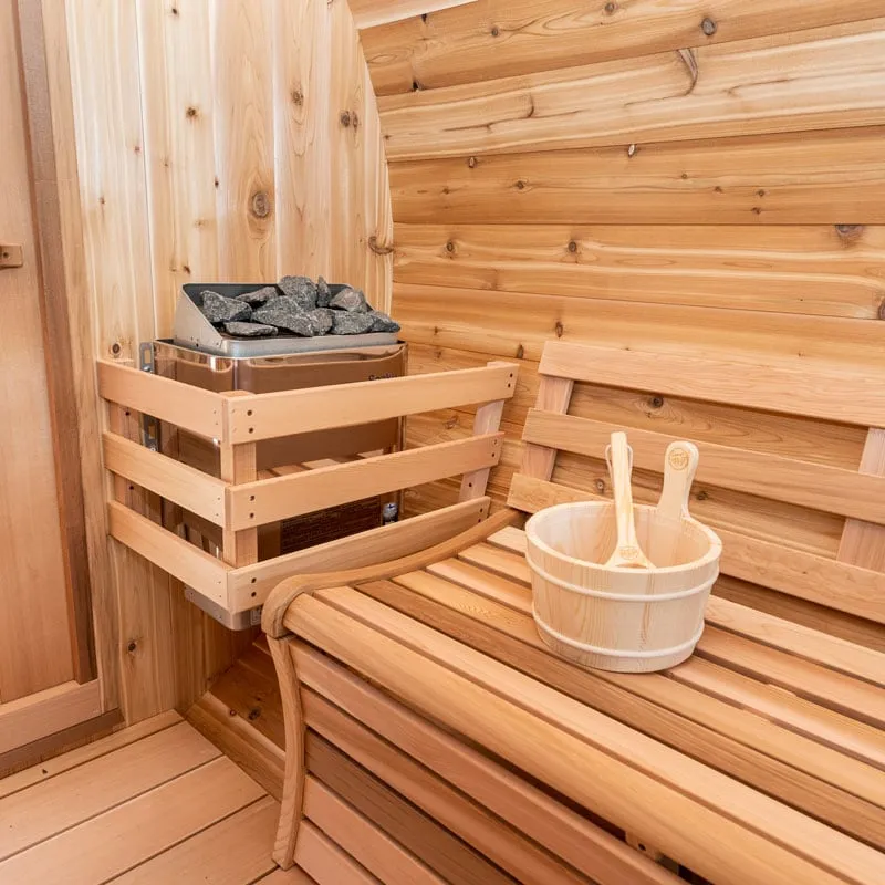 Barrel sauna interior corner: electric heater with rocks inside a timber guard, cedar bench with backrest, bucket and ladles