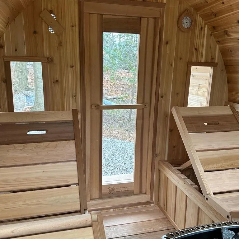 Barrel sauna interior facing the entry: glass-panel door, two small side windows, fold-up bench backrests and thermometer