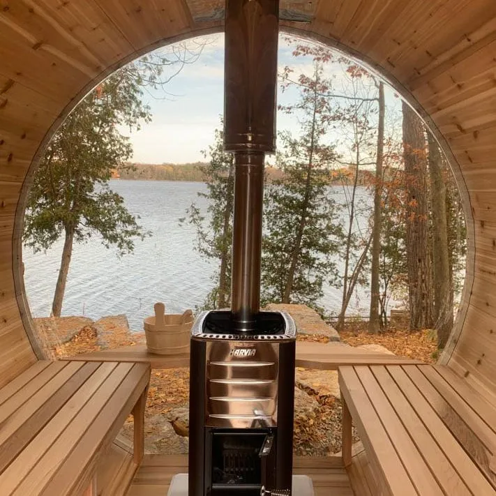 Barrel sauna interior with wood-burning stove and stainless flue in front of round glass end, cedar benches, autumn lake view