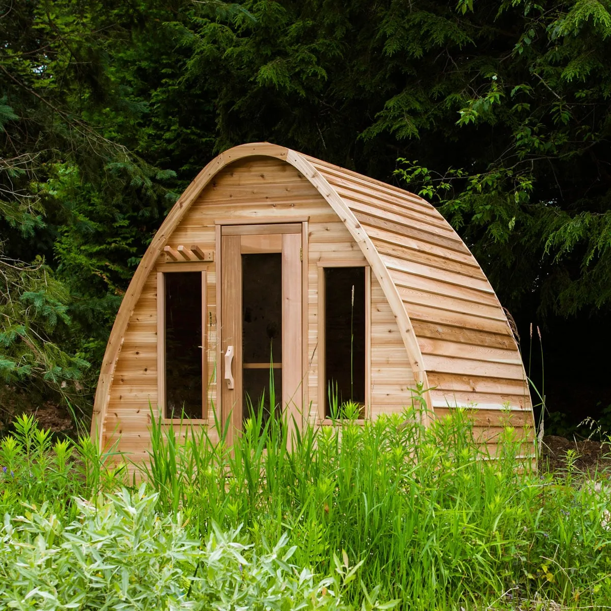 Arched pod sauna in knotty cedar with glass door, two front windows and towel pegs, in tall grass before evergreens