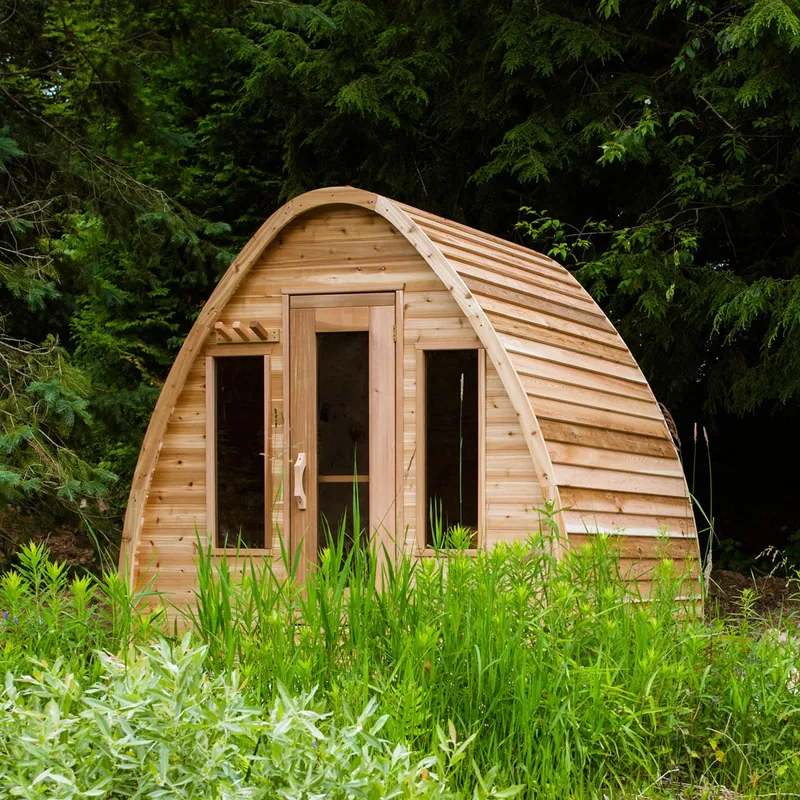 Arched pod sauna in knotty cedar with glass door, two front windows and towel pegs, in tall grass before evergreens