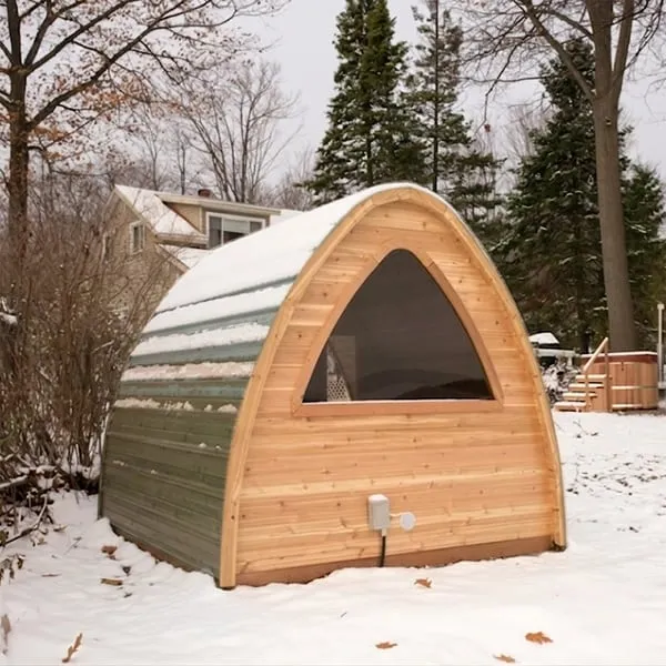 Rear of a cedar pod sauna with large arched window and green metal roof cladding under snow, in a snowy backyard
