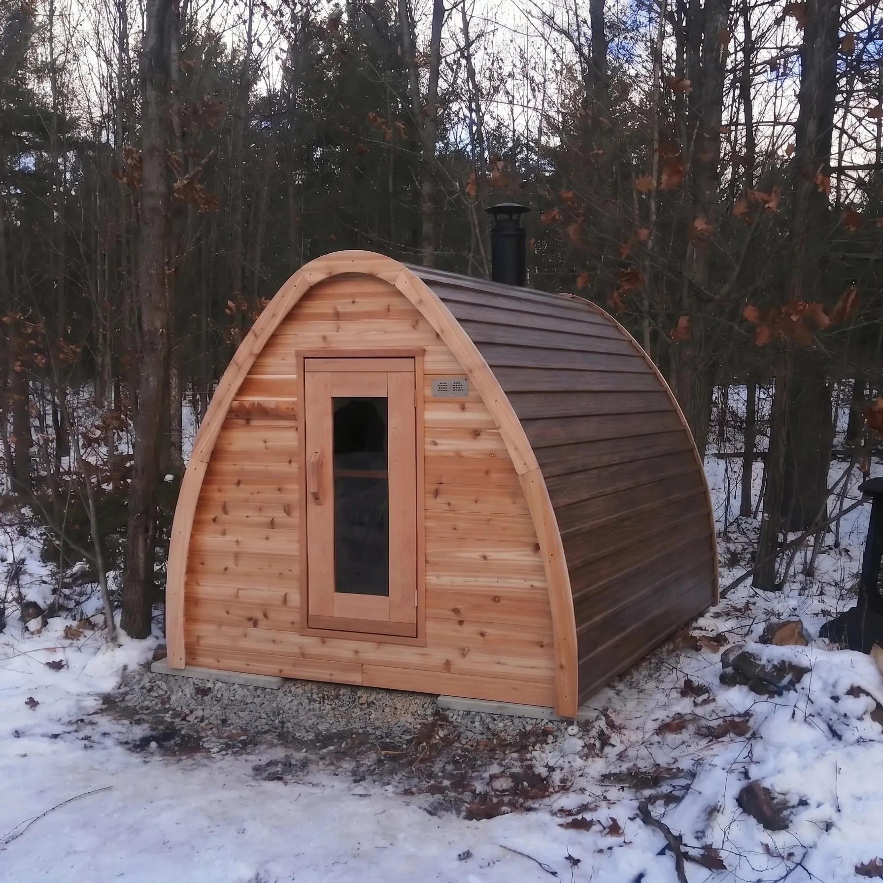 Cedar pod sauna with glass door, dark brown roof cladding and black chimney on a gravel pad in snowy woods