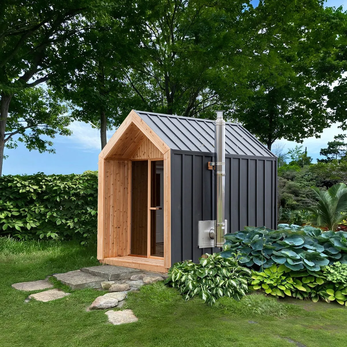 Gable-roof outdoor sauna with black metal siding and roof, cedar-lined front porch and glass door, stainless chimney, in a garden with stepping stones