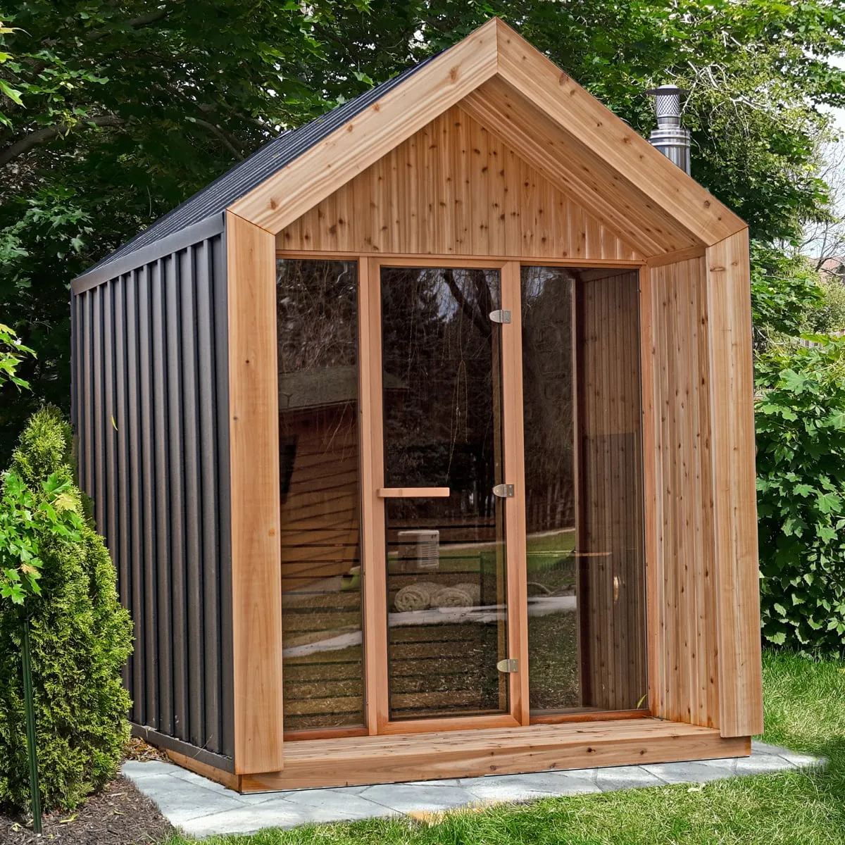 Gable-roof sauna with black metal siding and knotty cedar porch frame, full-width glass front with door, on pavers in a garden