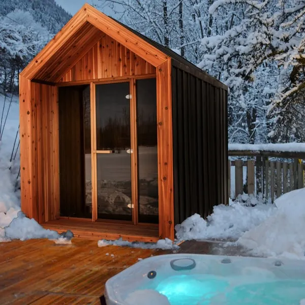 Gable-roof sauna with black siding and cedar porch on a snowy deck at dusk, hot tub in the foreground and snow-covered trees