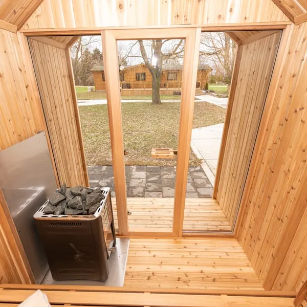 View from inside a knotty cedar sauna through the glass front and porch to a lawn, with a black stone-topped heater and metal heat shield