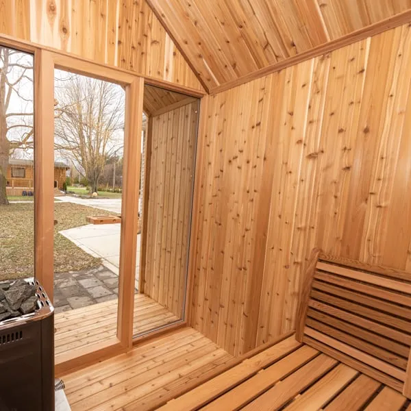 Knotty cedar sauna interior with glass front, open door to the porch, bench with backrest and edge of a black stone heater