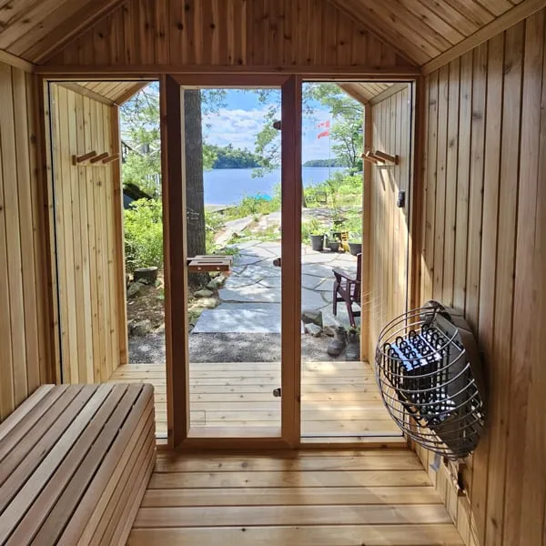 View from inside a cedar sauna through the glass door and porch to a lake, with a wall-mounted caged electric heater and bench