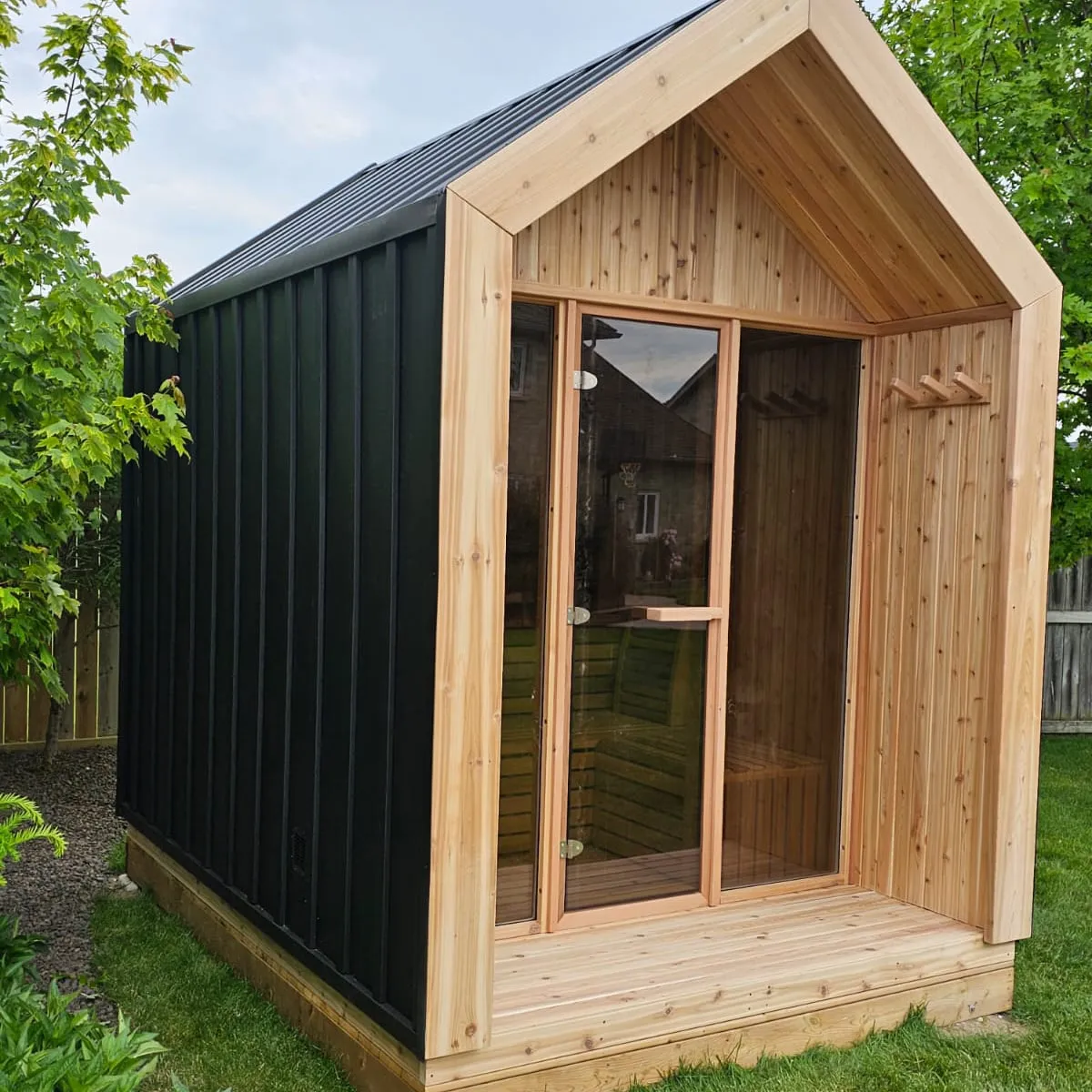 Gable-roof sauna with black metal siding, knotty cedar porch with towel pegs and full glass front, on a timber base in a backyard