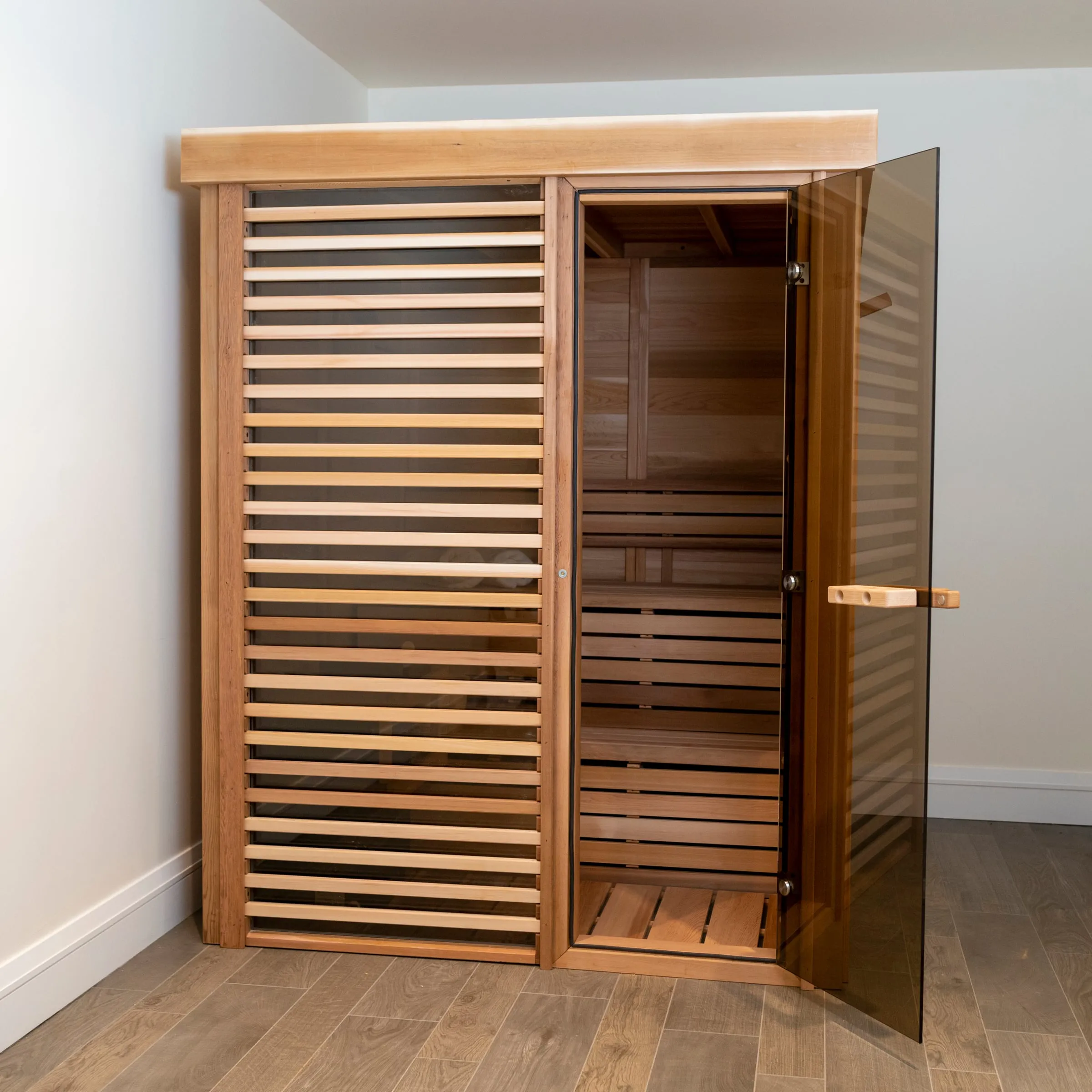 Indoor cedar cube sauna with horizontal louvres over the glass front panel and open bronze-tinted glass door, in a white-walled room