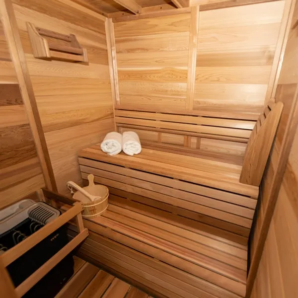 Clear cedar sauna interior with two-tier benches, backrest, rolled towels, bucket and ladle, and electric heater in a timber guard