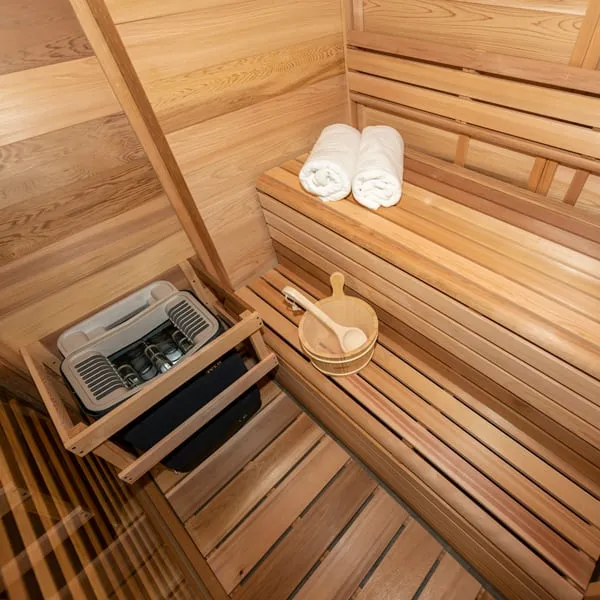 Overhead view of clear cedar sauna benches with rolled towels, bucket and ladle beside an electric heater in a timber guard